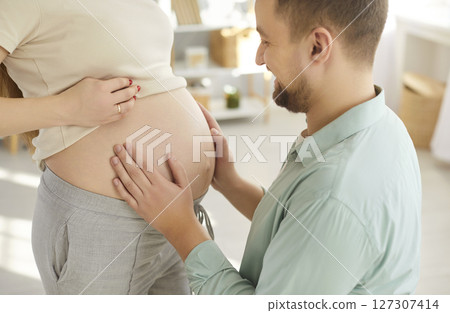 Closeup portrait of happy man looking at his beautiful pregnant woman's tummy sanding at home 127307414