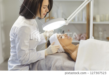 Cosmetologist looking, female facial treatment in spa salon, using professional magnifying lamp  127307523