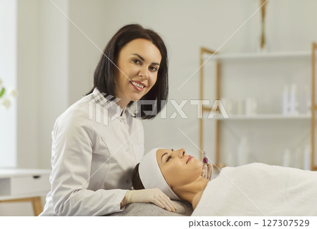 Cosmetologist looking at camera going to perform apparatus facial treatment procedure 127307529