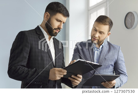 Portrait of two business men standing in office looking through financial documents. 127307530