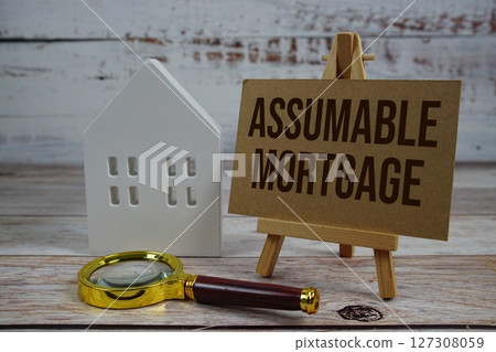 Assumable Mortgage on paper card with magnifying glass and white house model on wooden background 127308059