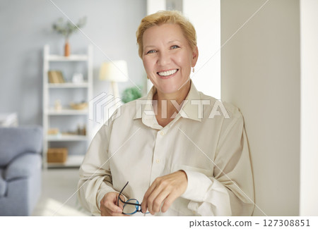 Portrait of mature woman standing in the living room and holding glasses in hand. Portrait of mature woman standing in the living room and holding glasses in hand. 127308851