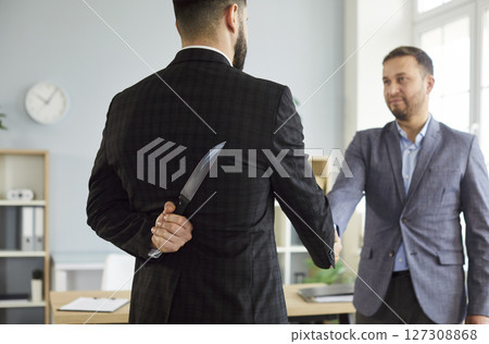 Businessman holding knife behind back and shaking hands with partner wearing suit in office. 127308868