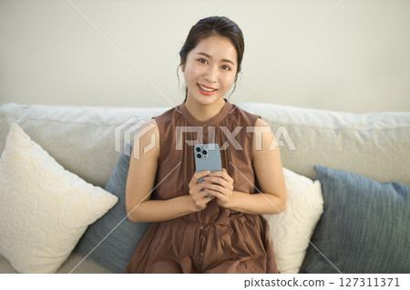 一位二十多歲的女性坐在沙發上操作智慧型手機 127311371