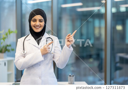 A smiling female doctor wearing a hijab and pointing towards an area in a modern medical office setting. She appears confident. 127311873