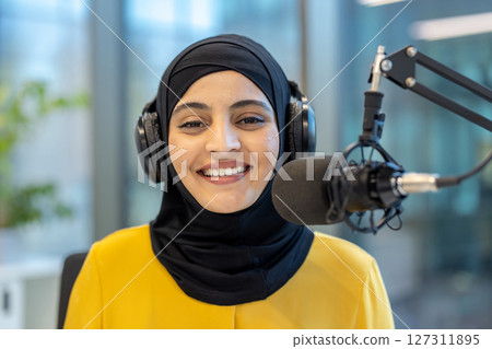 A young, smiling woman wearing a hijab and headphones speaks into a microphone, likely recording a podcast or radio show. 127311895