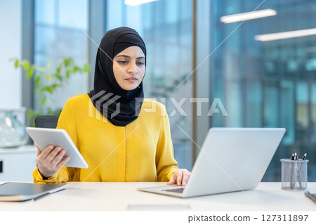 A professional woman in a yellow blouse and black hijab works at a desk with a laptop and tablet, possibly in an office setting. 127311897