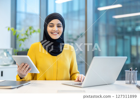A smiling woman wearing a hijab works at her desk in a modern office setting. She uses a tablet and a laptop, looking content. A smiling woman wearing a hijab works at her desk in a modern office setting. She uses a tablet and a laptop, looking content. 127311899