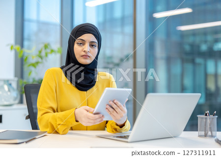 An Arab businesswoman in a yellow shirt and hijab works in an office, holding a tablet and using a laptop. An Arab businesswoman in a yellow shirt and hijab works in an office, holding a tablet and using a laptop. 127311911