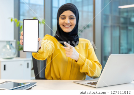 A smiling woman in hijab points to a blank smartphone screen, showcasing a modern workplace environment. 127311912