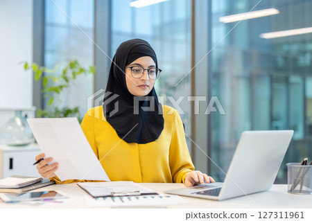 A professional woman wearing a hijab sits at her desk, reviewing documents and using a laptop, at the office. A professional woman wearing a hijab sits at her desk, reviewing documents and using a laptop, at the office. 127311961