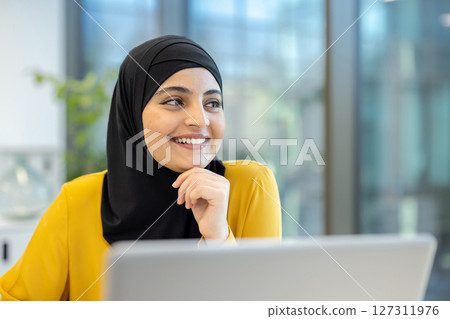 A smiling woman in a hijab is looking off-camera with her hand on her chin. She wears a yellow blouse, and a laptop is in front of her. 127311976