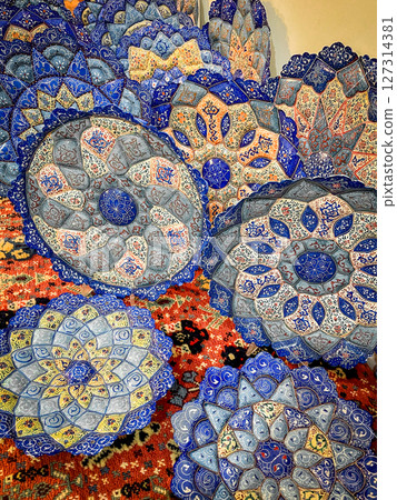 Intricately patterned Persian ceramic plates displayed on a vibrant rug, featuring hand-painted floral motifs in rich blue, orange, and grey tones 127314381