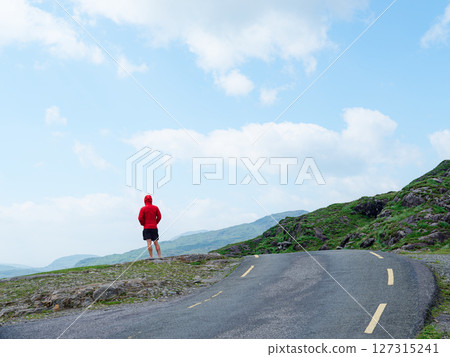 A person with a red hooded sweatshirt is standing on a rocky outcrop and looking out towards the scenic views of the Gap of Dunloe in County Kerry, Ireland. A person with a red hooded sweatshirt is standing on a rocky outcrop and looking out towards the scenic views of the Gap of Dunloe in County Kerry, Ireland. 127315241