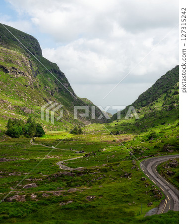 Breathtaking vista. winding road carves path through vibrant green Irish landscape, mountains framing valley. Breathtaking vista. winding road carves path through vibrant green Irish landscape, mountains framing valley. 127315242