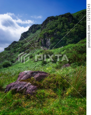 Strickeen mountain face covered in lush greenery and rocks on a bright, partly cloudy day in County Kerry, Ireland. Strickeen mountain face covered in lush greenery and rocks on a bright, partly cloudy day in County Kerry, Ireland. 127315243