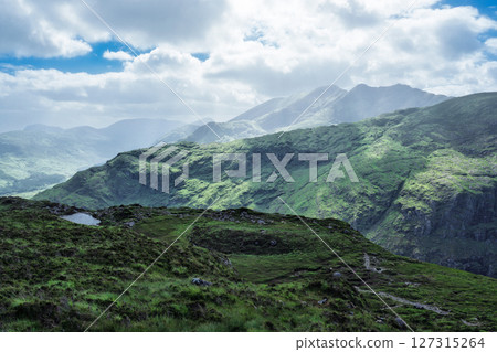 Rolling green hills lead to misty mountains in the distance, observed from the Head of the Gap of Dunloe. Soft light covers the landscape in County Kerry, Ireland. 127315264