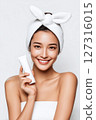 Smiling Asian woman with flawless skin holding a white skincare tube, wearing a towel wrap and a cute bow headband 127316015