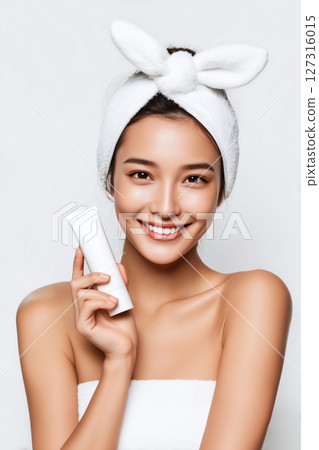 Smiling Asian woman with flawless skin holding a white skincare tube, wearing a towel wrap and a cute bow headband 127316015