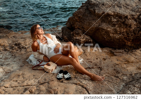 Woman Beach Relaxing - A woman in a white swimsuit and a white coverup relaxes on a beach with a rocky shore and ocean in the background. 127316962