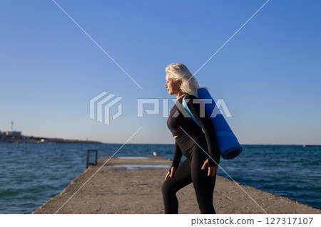 Woman Yoga Mat Pier - Senior woman in black leggings with blue yoga mat walking on pier overlooking the ocean. 127317107