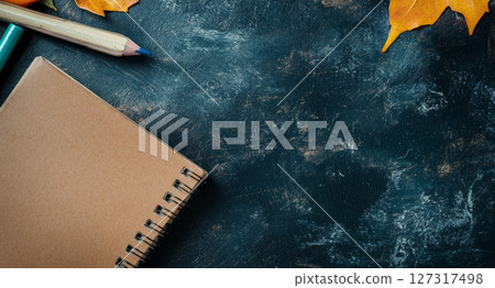 A brown notebook rests on a dark textured surface surrounded by colorful pencils and autumn leaves, perfect for back-to-school preparations 127317498