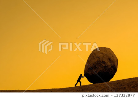 Silhouette of a Person Pushing a Boulder on a Mountain Slope at Dawn Against a Golden Sky 127322977