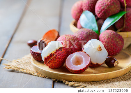 fresh ripe lychees in a bamboo basket with green leaves on a rustic wooden table. One peeled lychee reveals the juicy white flesh inside. 127323996