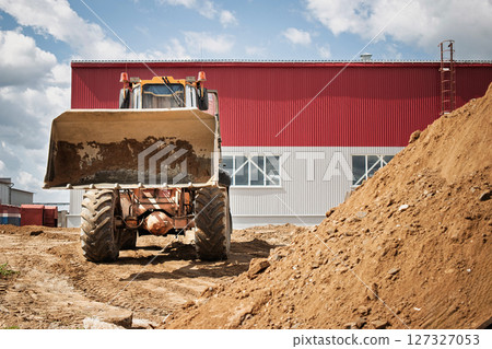 Heavy machinery is actively moving dirt on a construction site, with a large red and gray industrial building visible in the background. 127327053
