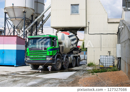 A cement truck waits while concrete is loaded at a construction site surrounded by industrial buildings in broad daylight. 127327086