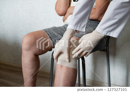 Medical professional treats a knee injury in a clinic setting with patient sitting in a chair 127327101