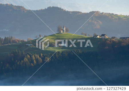 Beautiful autumnal landscape with forest, fog and small church on top a mountain Slovenia, Europe 127327429