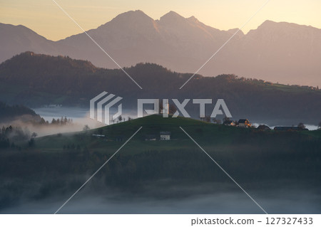 Landscape with church on top of a mountain surrounded by fog shot during colorful sunrise, Slovenia 127327433