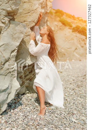 A woman in a white dress is standing on a rocky beach. Concept of serenity and tranquility, as the woman is enjoying the natural beauty of the beach. 127327462
