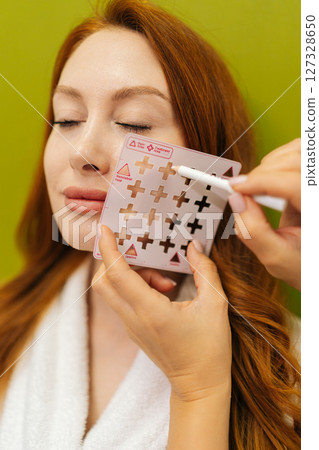 Vertical shot of cosmetologist holding markup grid on face of red-haired woman and marking with white pencil, preparing for beauty treatment on green background. Concept of beauty treatment. Vertical shot of cosmetologist holding markup grid on face of red-haired woman and marking with white pencil, preparing for beauty treatment on green background. Concept of beauty treatment. 127328650