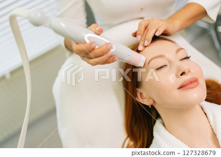 Close-up cropped shot of unrecognizable beautician using modern device performing mesotherapy procedure on forehead of young woman lying on treatment table in modern beauty salon. 127328679
