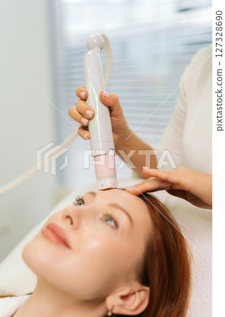 Close-up vertical shot of dermatologist using modern device performing mesotherapy procedure on forehead of woman lying on treatment table in beauty salon. Concept of anti-age rejuvenation skincare 127328690