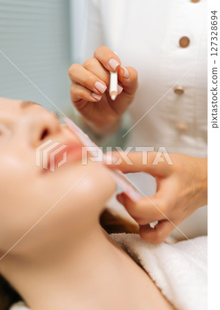 Vertical cropped shot of beautician preparing female client to facial cosmetic procedure, marking skin with white pencil and stencil, woman lying on treatment couch in beauty salon, close-up. Vertical cropped shot of beautician preparing female client to facial cosmetic procedure, marking skin with white pencil and stencil, woman lying on treatment couch in beauty salon, close-up. 127328694