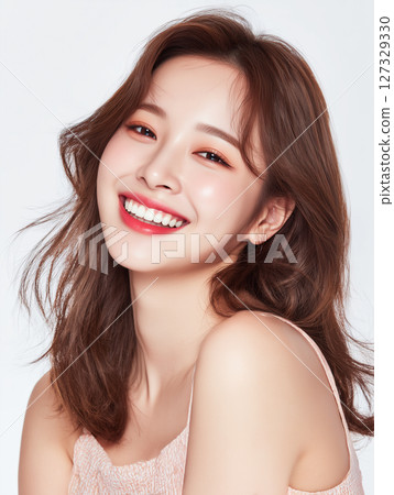 Smiling Korean woman with soft makeup and wavy hair in a light-colored outfit against a plain background 127329330