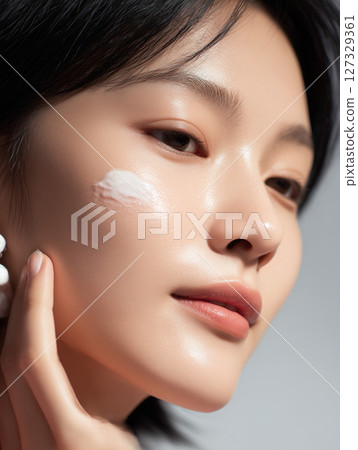 Korean woman applying skincare product while posing with a serene expression in a softly lit indoor setting 127329361
