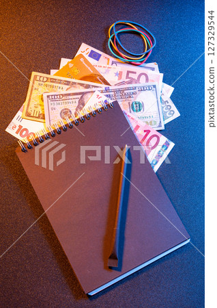 Assorted world currency with black notebook and pencil on dark background. 127329544