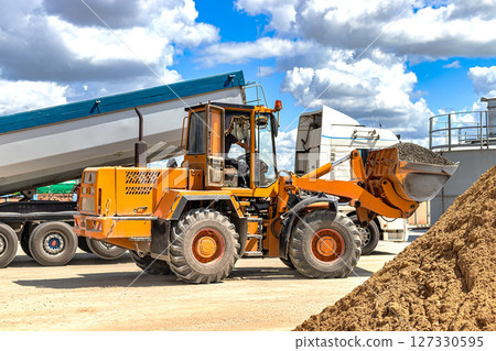 A large front loader transports crushed stone or gravel in a bucket at a construction site or concrete plant. Transportation of bulk materials. Construction equipment. Bulk cargo transportation. 127330595