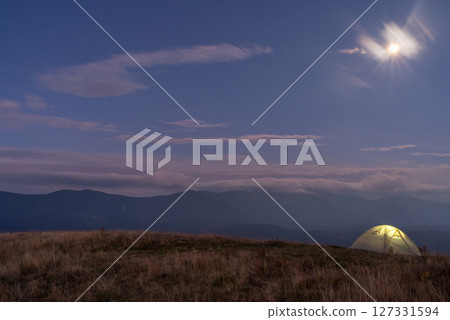 Beautiful mountain landscape. Tourist tent against the backdrop of mountains. Light in the tent that stands on the top of the mountain. 127331594