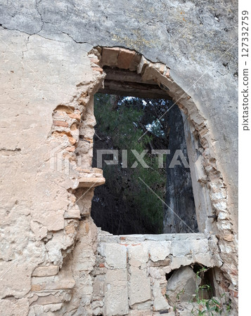 An opening in the crumbling wall of a historic stone structure in Spain reveals a glimpse of lush greenery outside. The remnants speak of a past long forgotten. 127332759