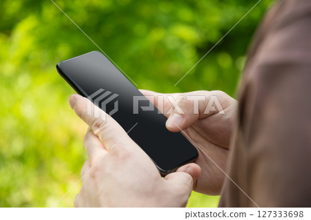 Close up of mans hands holding smartphone in outdoor green setting. Check apps, focused on device, chatting, texting. Fitness progress on app. Side view. Close up. 127333698