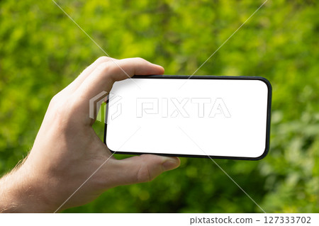 Man hand holding mobile phone with white blank screen against blurred green background. Showcasing mobile apps, design mockups, or digital content presentations. Check apps. Summer heat weather. Man hand holding mobile phone with white blank screen against blurred green background. Showcasing mobile apps, design mockups, or digital content presentations. Check apps. Summer heat weather. 127333702