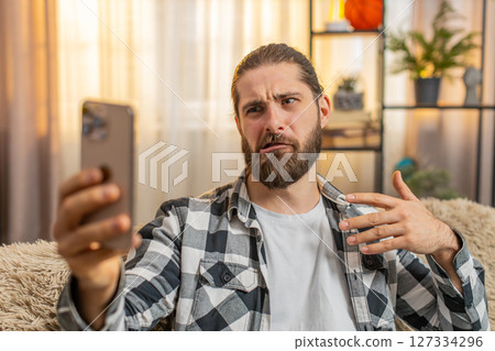 Young man blogger at home, sofa selfie mode, waving to camera, fun story, casual influencer vibe 127334296