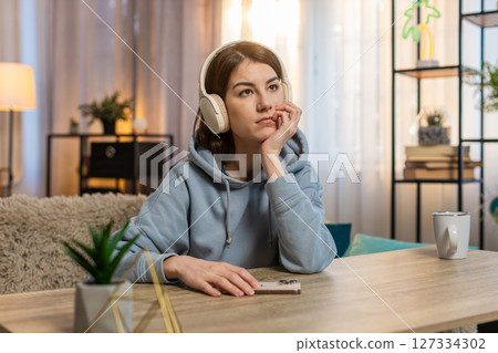 Young Caucasian woman listening to audiobook on smartphone with wireless headphones sitting on sofa 127334302