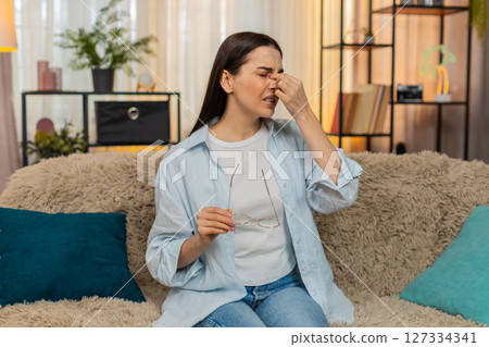 Young woman on sofa experiencing eye pain discomfort removing glasses and rubbing eyes tiredly 127334341