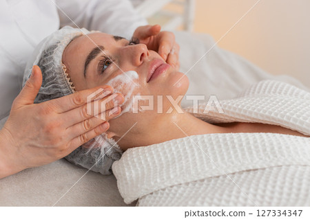 Cosmetologist gently massages woman face with foam cleanser during skincare procedure in spa clinic Cosmetologist gently massages woman face with foam cleanser during skincare procedure in spa clinic 127334347